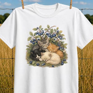 Op de afbeelding: Een wit t-shirt met korte mouwen voorzien van een grafische illustratie van vier kittens die samen onder een bosbessenstruik slapen. De kittens zijn zwart, grijs gestreept, oranje en crèmekleurig, genesteld tussen groene bladeren en blauwe bessen.