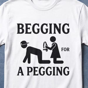 Op de afbeelding: Wit T-shirt met de zwarte tekst "BEGGING FOR A PEGGING". Het ontwerp toont stokfiguren. Het shirt heeft ook de tekst "100% COTTON WHITE T-SHIRT".
