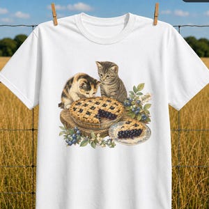 Op de afbeelding: Een wit t-shirt met een grafische illustratie van twee kittens die naar een bosbessentaart op een houten plank kijken. De ene kitten is lapjeskat en de andere is cyperse. Een stuk taart op een klein bordje en bosbessentakjes zijn afgebeeld.