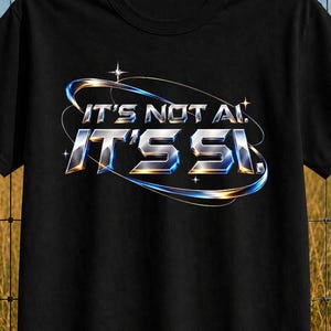 May include: A black short-sleeved t-shirt featuring a metallic, chrome-style graphic print on the chest. The text reads "IT'S NOT AI, IT'S SI." in a bold, stylised font, surrounded by a thin, glowing orbital ring design with small star accents.