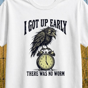 Könnte beinhalten: Ein weißes Kurzarm-T-Shirt mit einer Grafik eines schwarzen Raben, der auf einem Wecker sitzt. Der Text über dem Raben lautet I GOT UP EARLY und der Text unter der Uhr lautet THERE WAS NO WORM in einer dunkelblauen, fetten Schrift.