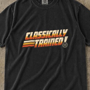 May include: A black short-sleeve t-shirt featuring the text "CLASSICALLY TRAINED" in a retro-style font with horizontal stripes in yellow, orange, and red. A small circular manual transmission gear shift pattern icon is located to the right.