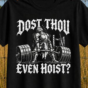 Op de afbeelding: Een zwart t-shirt met korte mouwen en een witte afbeelding van een ridder in harnas die een zware halterstang optilt. De tekst DOST THOU EVEN HOIST? is in een gotisch lettertype boven en onder de illustratie gedrukt.
