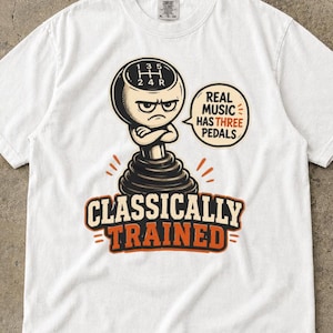 May include: A white short-sleeve t-shirt featuring a graphic of a manual gear shifter character with a grumpy expression. The text reads "REAL MUSIC HAS THREE PEDALS" and "CLASSICALLY TRAINED" in black and orange lettering on the front.
