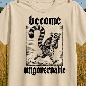 May include: A sand-colored t-shirt featuring a black ink-style illustration of a lemur running while holding a banana. The text above the lemur reads become and the text below reads ungovernable, both in a bold, gothic-inspired black typeface.
