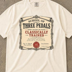 May include: An ivory short-sleeve t-shirt featuring a graphic print of a diploma-style border with the text "SCHOOL OF THREE PEDALS," "CLASSICALLY TRAINED," and a red manual transmission gear shift pattern icon in the center of the design.
