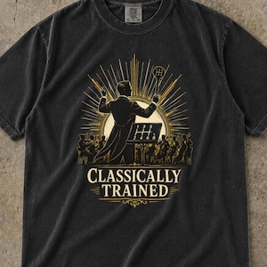 May include: A black short-sleeve t-shirt featuring a graphic of a conductor leading an orchestra with a manual gear shift knob as a baton. The text "CLASSICALLY TRAINED" appears below the illustration, which includes a gear shift pattern.