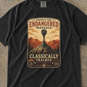 May include: A black short-sleeve t-shirt featuring a graphic print of a manual car gear shifter atop a mountain peak. The text reads: PROTECT THE ENDANGERED MANUALS, CLASSICALLY TRAINED, SINCE FOREVER, SAVE SHIFT, and 6 SPEED SERIES.