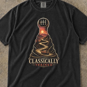 Może przedstawiać: Czarny t-shirt z krótkim rękawem z grafiką przedstawiającą gałkę manualnej skrzyni biegów, wewnątrz której widać krętą górską drogę o zachodzie słońca z samochodem. Poniżej znajduje się napis "CLASSICALLY TRAINED" w kremowym kolorze.