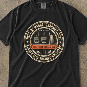 May include: A black short-sleeve t-shirt featuring a circular graphic with the text DEPT. OF MANUAL TRANSMISSIONS CLASSICALLY TRAINED OPERATOR and EST. THREE PEDALS AGO, along with illustrations of three car pedals and a gear shift pattern.