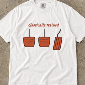 Puede incluir: Una camiseta blanca de manga corta con un gráfico de tres pedales de coche en color naranja con contornos negros. Sobre los pedales, el texto "classically trained" está impreso en una fuente de color naranja oscuro. La camiseta tiene cuello redondo.