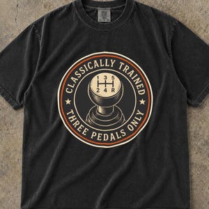 May include: A black short-sleeve t-shirt featuring a circular graphic on the chest. The design depicts a manual gear shifter with a five-speed shift pattern and the text "CLASSICALLY TRAINED" and "THREE PEDALS ONLY" in a circular border.