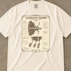 Puede incluir: Una camiseta de manga corta color marfil con un estampado gráfico marrón oscuro de un dibujo técnico de patente titulado Classically Trained. El diseño muestra un mecanismo de embrague y tres pedales con etiquetas técnicas.