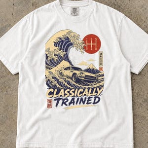May include: A white short-sleeve t-shirt featuring a graphic print of a vintage sports car driving through a large ocean wave, a red circle with a manual gear shift pattern, Mount Fuji, Japanese characters, and the text Classically Trained.