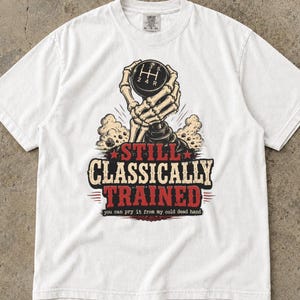 May include: A white short-sleeve t-shirt featuring a graphic of a skeleton hand gripping a manual gear shift knob. The text reads "STILL CLASSICALLY TRAINED" in red and black, with "you can pry it from my cold dead hand" printed below.