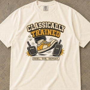 May include: An ivory short-sleeve t-shirt featuring a graphic of a foot in a yellow and white sneaker pressing a brake and gas pedal. The text reads "CLASSICALLY TRAINED" and "HEEL. TOE. REPEAT." The shirt is labeled as ivory and has a crew neckline.