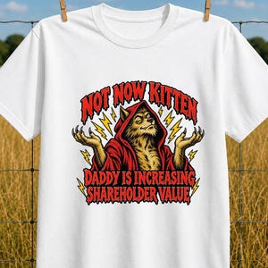 Könnte beinhalten: Ein weißes T-Shirt mit kurzen Ärmeln und einer Grafik eines Wolfs in einer roten Kapuzenrobe mit erhobenen Händen. Der Text NOT NOW KITTEN DADDY IS INCREASING SHAREHOLDER VALUE steht in roten Buchstaben, umgeben von gelben Blitzen.