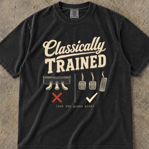 May include: A black short-sleeve t-shirt featuring the text Classically TRAINED in cream. Below, an illustration shows piano pedals with a red X and car pedals with a white checkmark, followed by the text (not the piano kind).