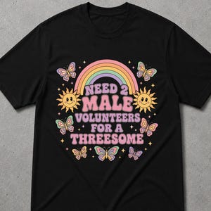 Könnte beinhalten: Ein schwarzes Kurzarm-T-Shirt mit einem farbenfrohen Grafikdruck. Das Design zeigt einen pastellfarbenen Regenbogen, zwei lächelnde Sonnen und mehrere Schmetterlinge um den Text "NEED 2 MALE VOLUNTEERS FOR A THREESOME" in einer fetten, rosa Retro-Schrift.