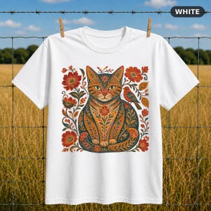 Könnte beinhalten: Ein weißes T-Shirt mit einer zentralen Grafik einer Katze, umgeben von komplizierten Blumenmustern und einem kleinen Vogel. Das Design verwendet eine Farbpalette aus Orange-, Rot- und Dunkelblautönen. Das Shirt hat kurze Ärmel und einen Rundhalsausschnitt.