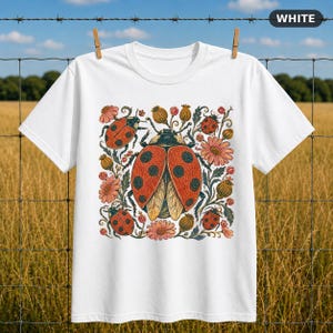 Può includere: Una t-shirt bianca a maniche corte con una stampa grafica centrale di una grande coccinella circondata da coccinelle più piccole, fiori rosa e baccelli. Il design utilizza uno stile illustrativo dettagliato con toni arancioni, rossi e verdi.