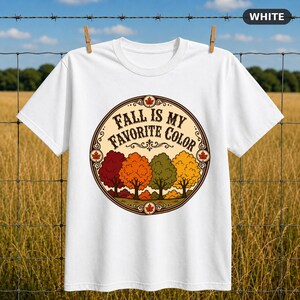 May include: A white short-sleeve t-shirt featuring a circular graphic with the text "FALL IS MY FAVORITE COLOR" in black lettering. The design includes a row of stylized trees in shades of red, orange, green, and yellow, accented by small maple leaf icons.