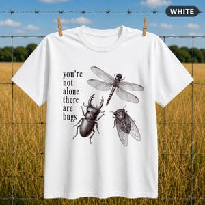 Può includere: Una t-shirt bianca a maniche corte con un'illustrazione a inchiostro nero di una libellula, un cervo volante e una cicala. Il testo "you're not alone there are bugs" è stampato in lettere nere a sinistra delle illustrazioni degli insetti.