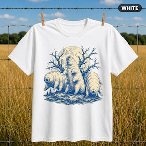 Può includere: Una t-shirt bianca a maniche corte con una stampa grafica blu di tre tardigradi in piedi su una superficie strutturata davanti a una grande luna e rami d'albero spogli. La maglietta presenta un girocollo.