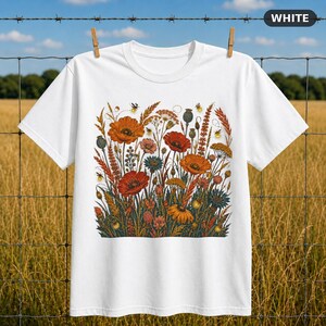 Peut inclure: Un t-shirt blanc à manches courtes avec une impression graphique centrale représentant une prairie de fleurs sauvages. L'illustration comprend des coquelicots orange, des épis de blé et divers petits insectes comme des lucioles.