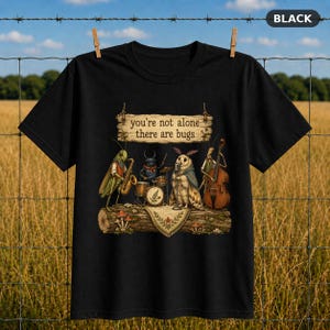 Può includere: Una t-shirt nera con un'illustrazione di insetti che suonano strumenti musicali su un tronco. Il testo recita, you're not alone there are bugs. L'immagine include una cavalletta con un sassofono, uno scarabeo alla batteria e una falena.