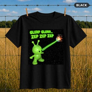 Op de afbeelding: Een zwart t-shirt met een afbeelding van een felgroene alien in de ruimte. De alien schiet een groene laserstraal naar een kleine planeet. Boven de alien staat de tekst GLERP GLORP... ZEP ZEP ZEP in een neon-groen lettertype.