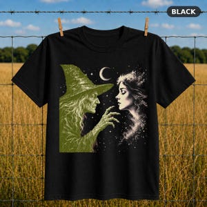 Op de afbeelding: Een zwart t-shirt met korte mouwen en een grafische print van een oude heks met een puntmuts die naar het gezicht van een jonge vrouw reikt. Het ontwerp bevat een maansikkel en sterren tegen een donkere achtergrond in groentinten.