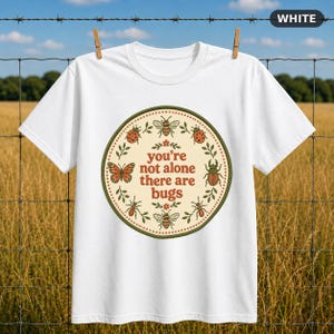 May include: A white short-sleeve t-shirt featuring a circular graphic with a cream background and a dark green border. The text "you're not alone there are bugs" is centered, surrounded by illustrations of a butterfly, bees, ants, and beetles.
