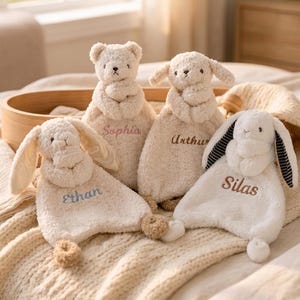 Pode incluir: Quatro mantas de segurança de pelúcia com cabeças e membros de animais. Um urso com o nome Sophia, um cão com o nome Arthur e dois coelhos com os nomes Ethan e Silas bordados com fio colorido no tecido macio de cor creme.