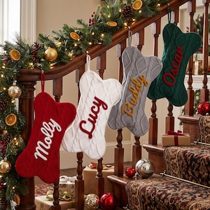 May include: Four bone-shaped Christmas stockings made of cable-knit fabric, hanging from a wooden staircase railing. The stockings are red, white, grey, and dark green, each featuring a name embroidered in a contrasting colour: Molly, Lucy, Buddy, and Oscar.
