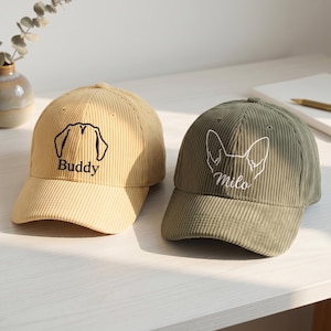 Può includere: Due cappellini da baseball in velluto a coste. Il cappellino a sinistra è color sabbia con un ricamo nero del profilo di una testa di cane e il nome Buddy. Quello a destra è verde oliva con un ricamo bianco del profilo di un cane e il nome Milo.