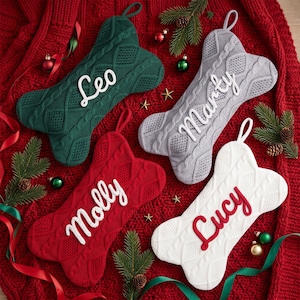 May include: Four bone-shaped Christmas stockings in dark green, light grey, red, and white. Each stocking features a cable knit texture and a hanging loop. The stockings are personalised with white or red script lettering reading Leo, Marty, Molly, and Lucy.