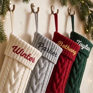 May include: Four cable-knit Christmas stockings hanging from metal hooks. The stockings are cream, light grey, red, and dark green. Each features a folded cuff embroidered with a name: Winter in red, Parker in black, Bethany in gold, and Taylor in white.