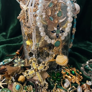 Può includere: Una collezione di gioielli vintage, tra cui collane, bracciali e spille, in una varietà di stili e materiali. I pezzi presentano perle, conchiglie e metallo dorato, con accenti di turchese e pietre preziose verdi. I gioielli sono esposti su una superficie di velluto verde scuro.