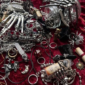 Puede incluir: Una colección de joyas y accesorios de temática gótica sobre una superficie de terciopelo rojo. Los artículos incluyen collares, anillos, colgantes y una mano de esqueleto plateada. Un tarro de cristal contiene más anillos.
