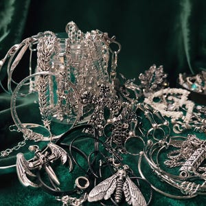 Puede incluir: Una variedad de joyas plateadas, que incluyen collares, pulseras y anillos, se exhibe sobre un fondo de terciopelo verde oscuro. Las joyas presentan diseños góticos y macabros, como esqueletos, cruces y colgantes de polilla. Las piezas son intrincadamente detalladas.