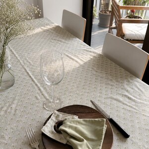 Peut inclure: Une table de salle à manger dressée pour un repas. La table est recouverte d'une nappe crème à motifs floraux discrets. Une assiette en bois contient une serviette verte pliée, un anneau de serviette et un couteau. Un verre à vin et une fourchette sont également sur la table.