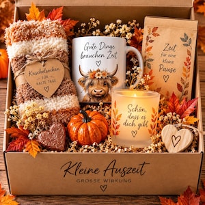 Könnte beinhalten: Eine Geschenkbox mit flauschigen Ringelsocken mit herzförmigem Anhänger, einer weißen Tasse mit Hochlandrind-Motiv und deutschem Text, einer Kerze mit Text, einer Papiertüte, einem kleinen Kürbis und einem herzförmigen Schokoladenstück.