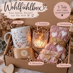 Könnte beinhalten: Eine Geschenkbox mit einer weißen Keramiktasse mit Blumenmotiven und dem Text 'Schön, dass es dich gibt', einem Teelichtglas mit schwarzer Linienkunst, flauschigen rosa-weißen Herzsocken sowie zwei verpackten Überraschungsartikeln.