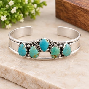 May include: A silver cuff bracelet featuring five teardrop-shaped turquoise stones arranged in a row. The stones vary in shades of blue and green with natural veining. The metal band has a smooth, polished finish with small silver bead accents.