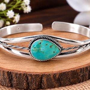 May include: A silver cuff bracelet featuring a central teardrop-shaped turquoise stone with brown matrix inclusions. The band is constructed from two smooth silver wires and one twisted silver wire, meeting at the stone setting with a rope border.