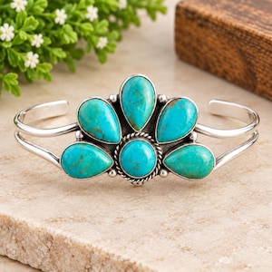 May include: A silver cuff bracelet featuring five teardrop-shaped turquoise stones and one central round turquoise stone. The stones are set in polished silver with a twisted rope detail surrounding the centre stone and a split band design.