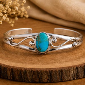 May include: A silver cuff bracelet featuring a central oval turquoise stone with brown matrix veining. The metal band displays a polished finish with decorative swirling wire accents on both sides of the stone, creating an open, split-shank design.