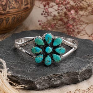 May include: A silver cuff bracelet featuring a central flower-shaped cluster of nine teardrop-shaped turquoise stones. The metal band splits into two arms on each side, connecting to the circular stone setting which has a dark oxidised finish.