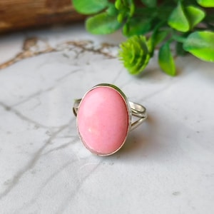 May include: A silver ring featuring a large, oval-shaped, light pink gemstone. The gemstone is set in a silver band with a split shank design. The ring is displayed on a white and gray marble surface.